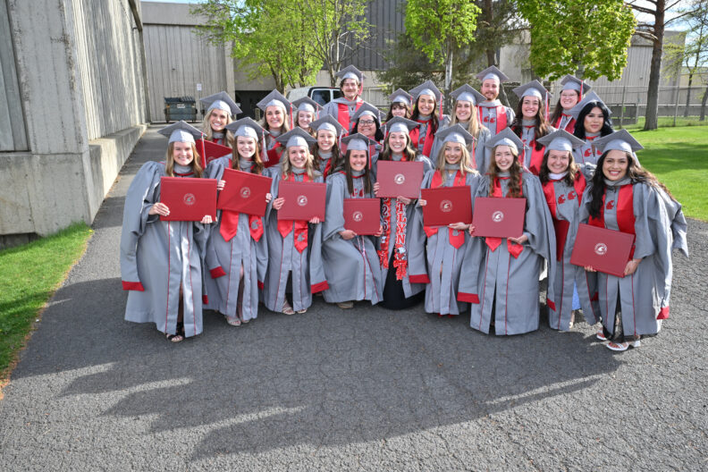 Group of graduates in gray gowns holding diplomas outdoors.