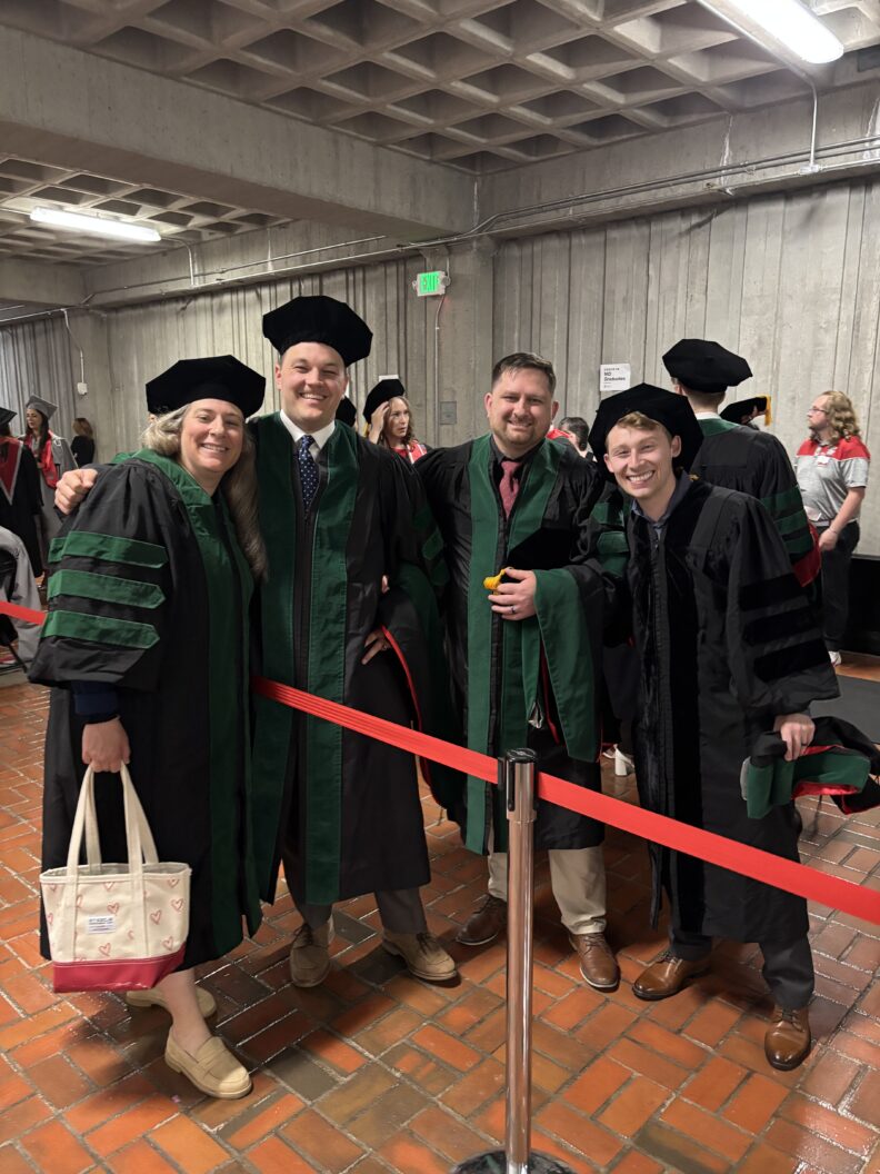 Four graduates in black and green regalia smiling near a roped-off area.