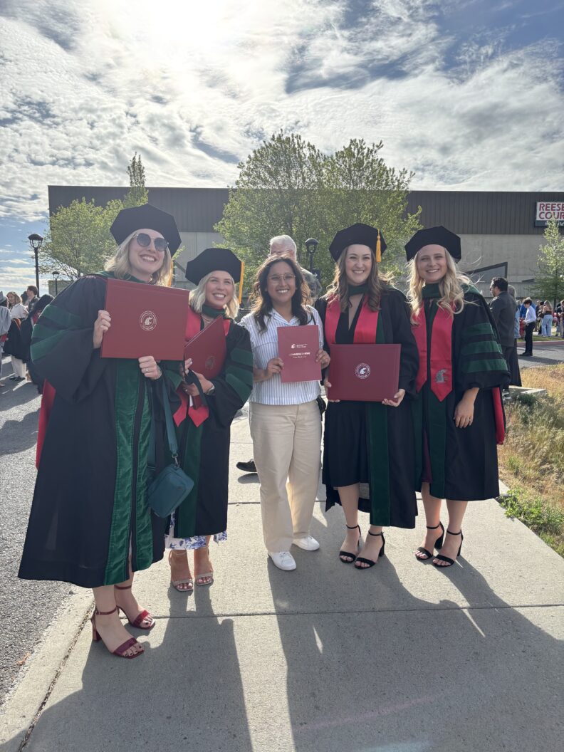 Graduates in regalia hold diplomas outdoors on a sunny day.
