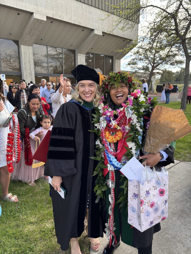 Two graduates pose outside, one wearing multiple leis and holding flowers.