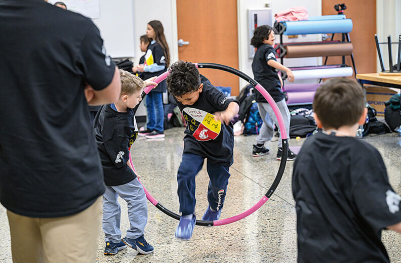 Children participating in a group activity indoors, with one child stepping through a colorful hoop while others watch and play nearby.