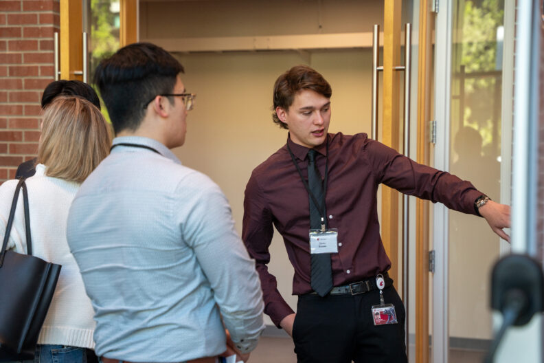 Student presenter gestures while speaking with attendees in a hallway.