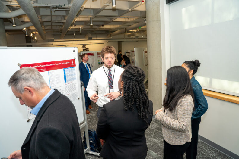 Student presenter discusses research with attendees beside a poster display.