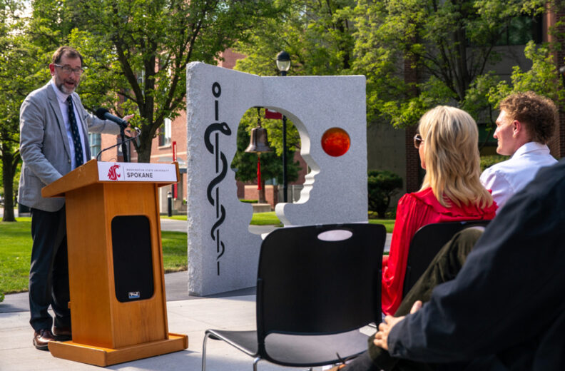 Speaker at a podium addresses an outdoor audience beside a medical-themed sculpture.