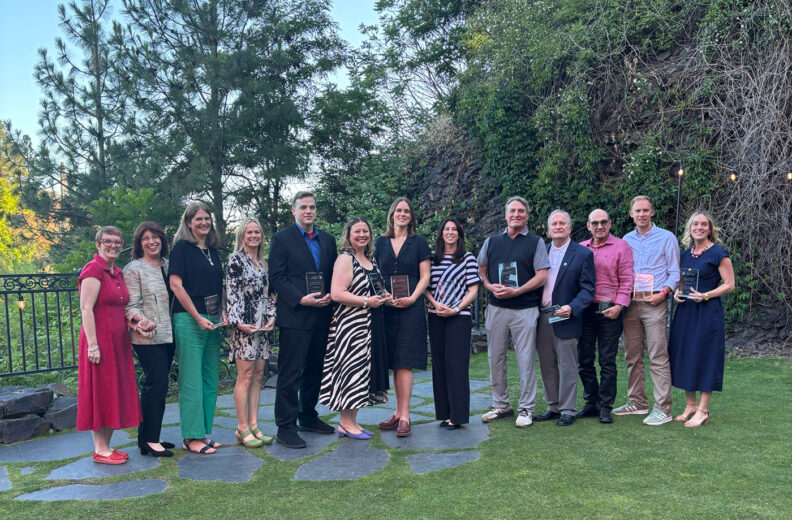 A group of adults stand in a line outdoors on a lawn and stone pathway, each holding a glass award plaque.