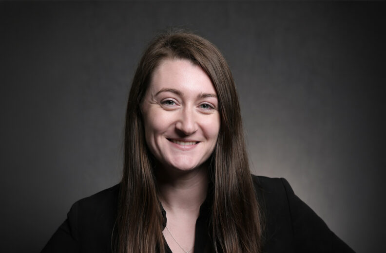 Professional headshot of a person in black attire against a dark gray background.