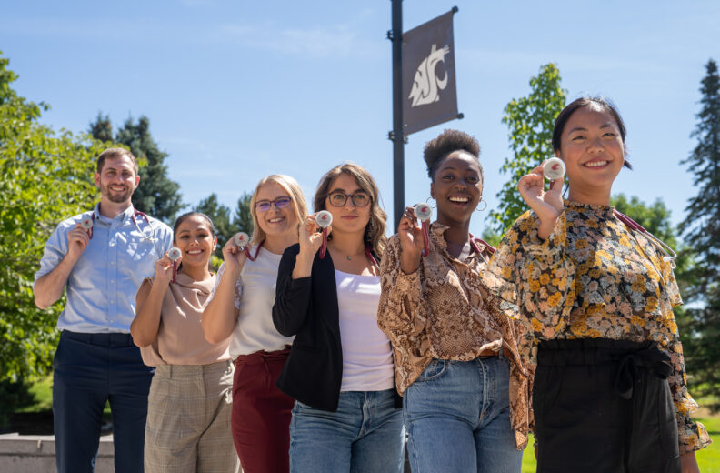 Six people pose outdoors holding stethoscopes, with a Washington State University banner and trees visible in the background.