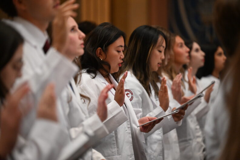 A line of students in their White Coats holding their hand up reciting the student declaration. 