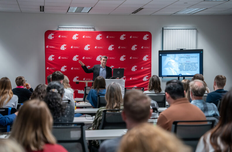 Presenter speaks to an audience in a classroom, standing at a podium in front of a red Washington State University College of Medicine backdrop and presentation screen.