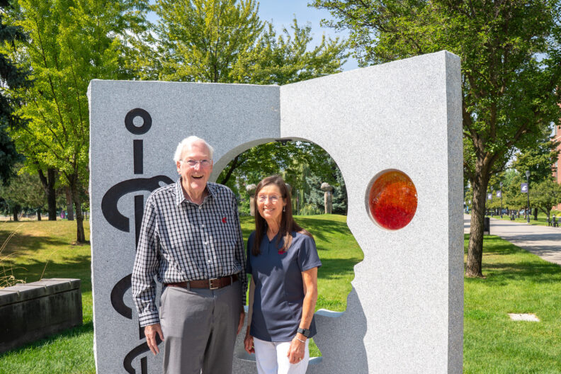 An elderly couple stands outside in front of a College of Medicine sculpture. 