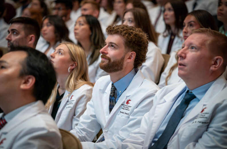 Medical students in white coats sit in an auditorium and listen during a White Coat Ceremony.