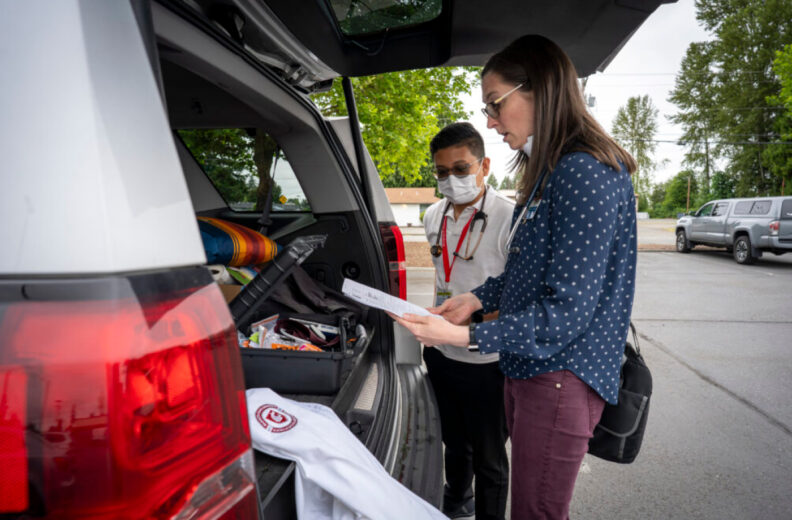 Two healthcare team members review a checklist beside an open vehicle filled with medical supplies and equipment in a parking lot.