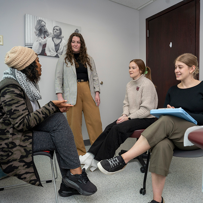 Standardized patient sitting and talking to two graduate students while professor stands and listens. 