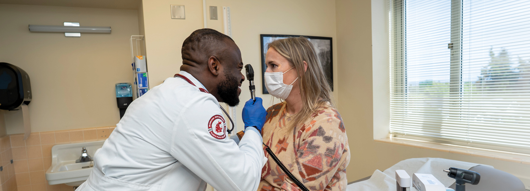 Resident using a tool to look into a patient's eye, inside an exam room at a clinic.