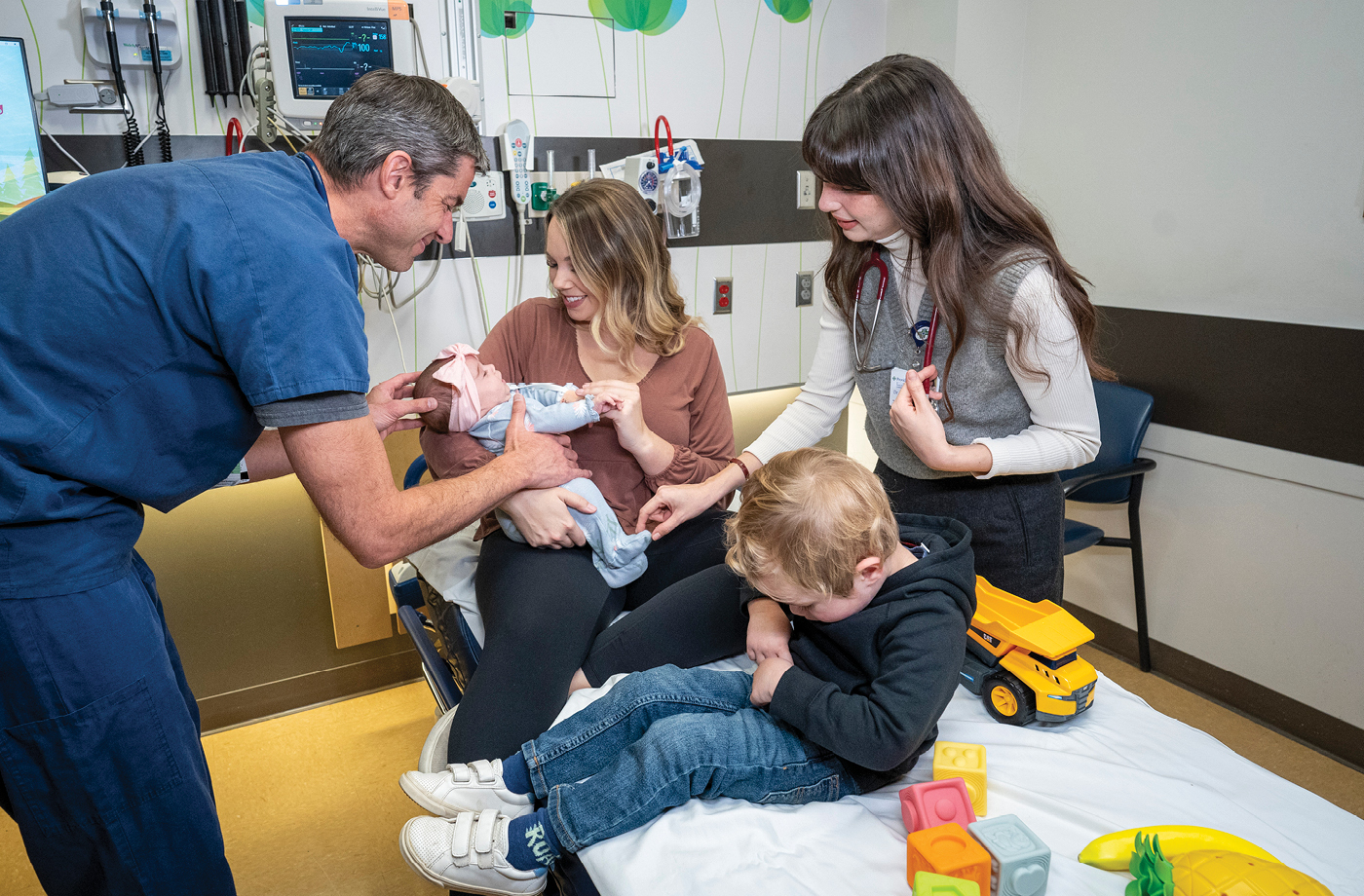 A doctor interacts with a baby being held by an adult seated on a hospital bed. A resident with a stethoscope stands nearby, observing. 