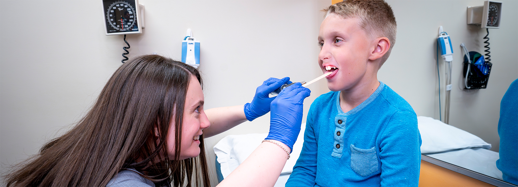 Pediatric resident looking in a child's mouth with a light in an exam room.