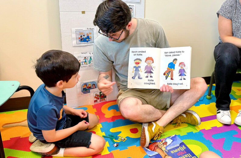 Student reading book with a child on the floor.