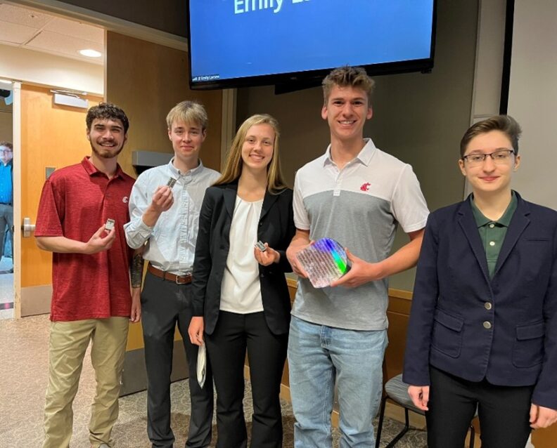 5 men and women posing in front of a video display along a wall.