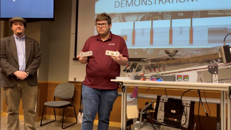 A man holding up an injection-molded part in both hands while standing in front of a large screen giving a presentation.