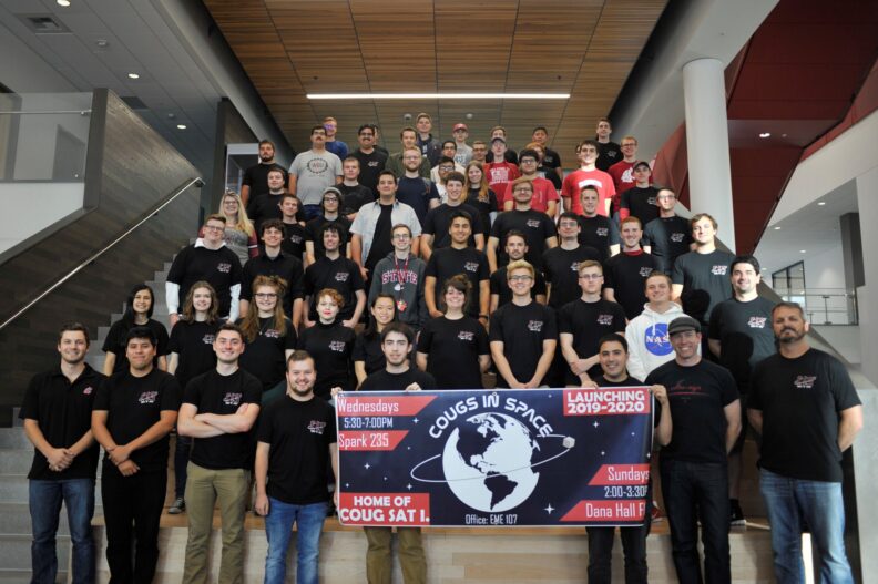 Large group of students standing on a wide indoor staircase, many wearing black shirts with CIS logo, posing together behind a banner that reads Cougs in Space.