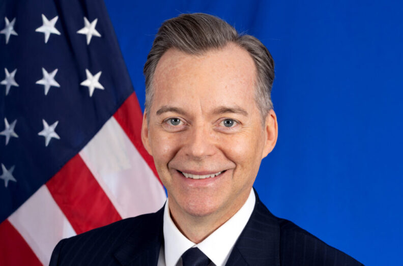 Ambassador Roger Nyhus in front of American flag.