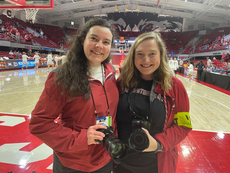 Two women hold cameras and pose for a picture on a basketball court