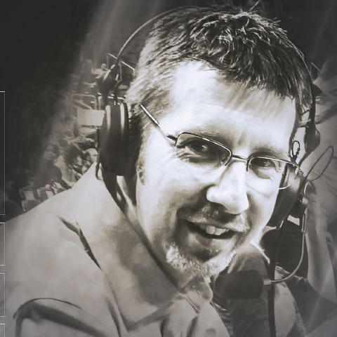 Black-and-white close-up portrait of a sports broadcaster wearing a headset with a microphone and glasses. The broadcaster looks toward the camera while seated at a broadcast position, with a blurred crowd and event setting visible in the background.