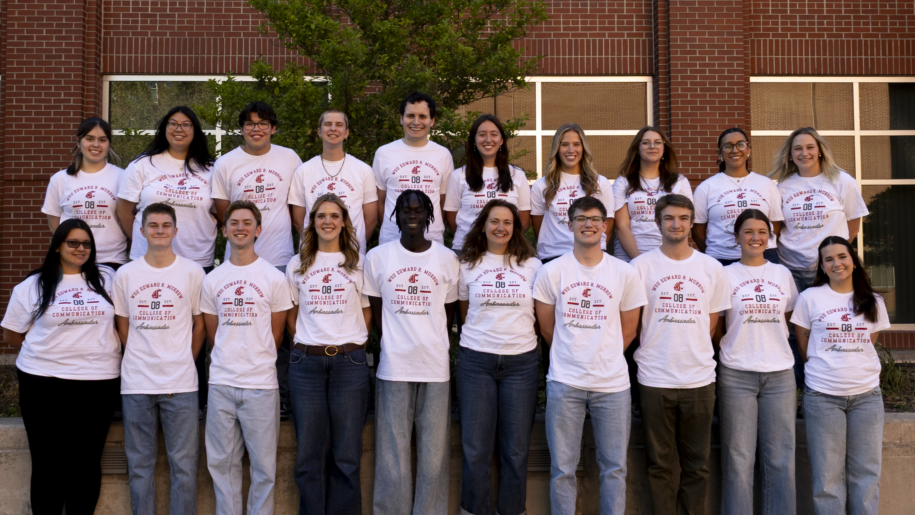 Group photo of 20 Murrow College of Communication ambassadors wearing white ambassador T-shirts, standing in two rows outdoors in front of a red-brick campus building with large windows and a tree.