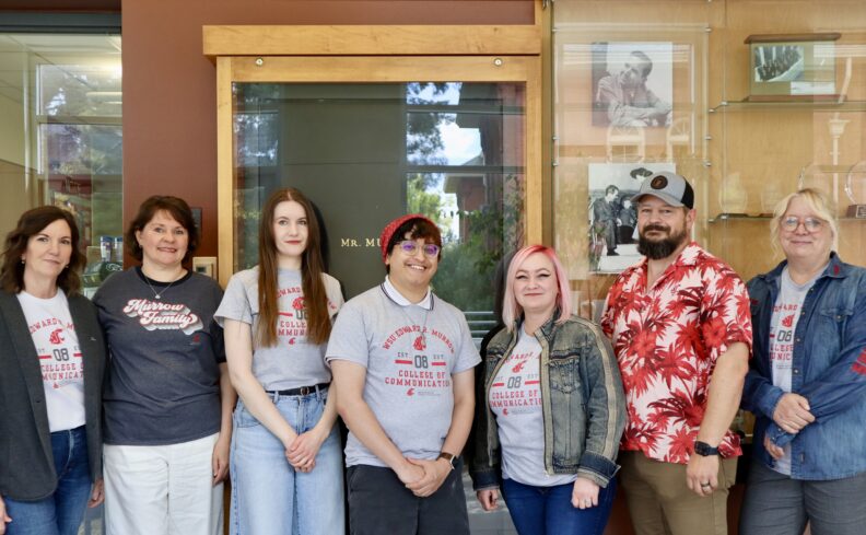 Seven people stand together indoors in front of a wooden display case and glass trophy cabinet. Several are wearing gray Edward R. Murrow College of Communication T-shirts, while others wear casual layers, including a denim jacket and a red floral shirt. Framed photographs and awards are visible in the background, and the group is posed for a portrait-style photo.
