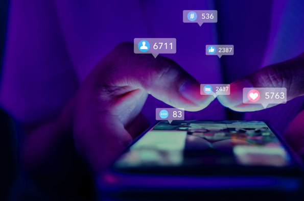 Close-up of a person’s hand interacting with a smartphone screen illuminated by purple and blue light. Floating social media notification icons and engagement metrics, including likes, comments and follower counts, appear above the device, illustrating online communication, social media activity and digital engagement.