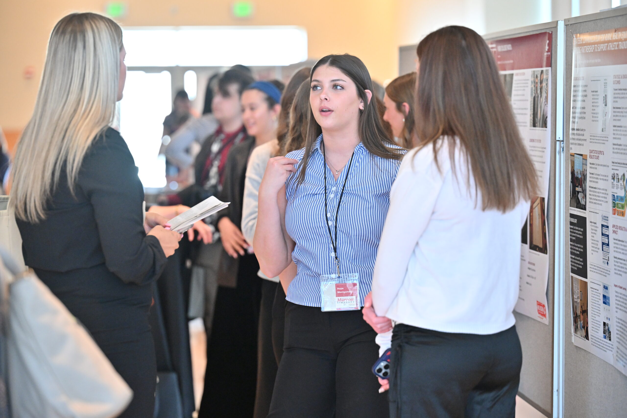 Students stand alongside research and project presentation posters at an academic showcase. In the foreground, three students in professional attire discuss a poster presentation, with one student wearing a conference name badge and gesturing while speaking. Additional students and attendees are visible in the background, viewing displays and engaging in conversations throughout the bright event space. Rows of poster boards featuring text, images and research findings line the room, highlighting student scholarship and professional communication skills.