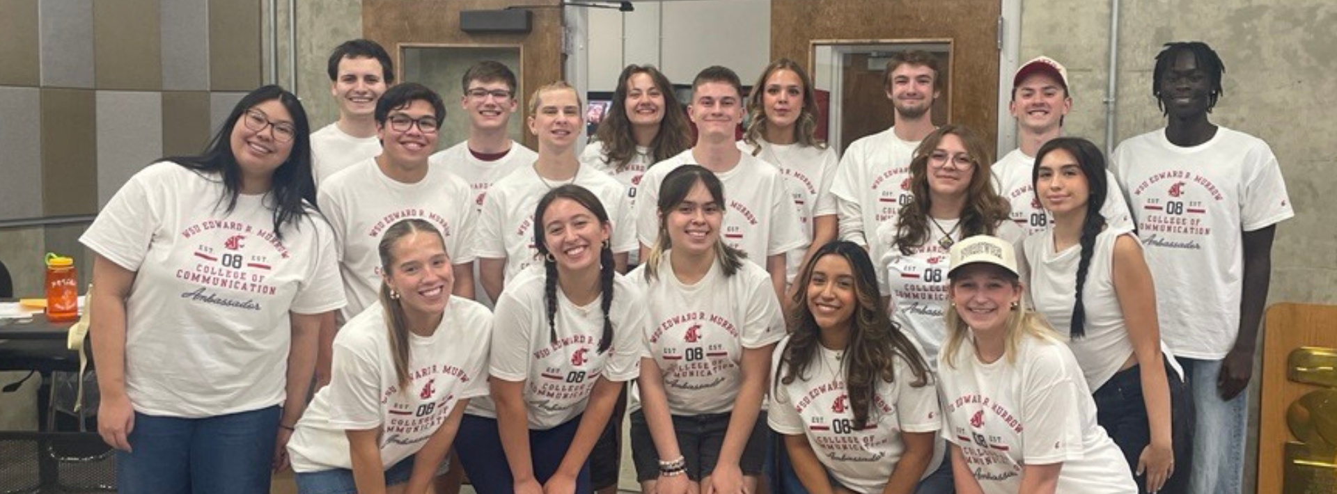 A group of approximately 15 students wearing matching Edward R. Murrow College of Communication T-shirts pose for a photo inside a media production or classroom space. The students stand in two rows in front of a concrete wall and double doors, with acoustic wall panels visible on the left. A large award plaque rests against the wall on the right side of the room. Tables, chairs and classroom equipment are visible in the background under bright overhead lighting.
