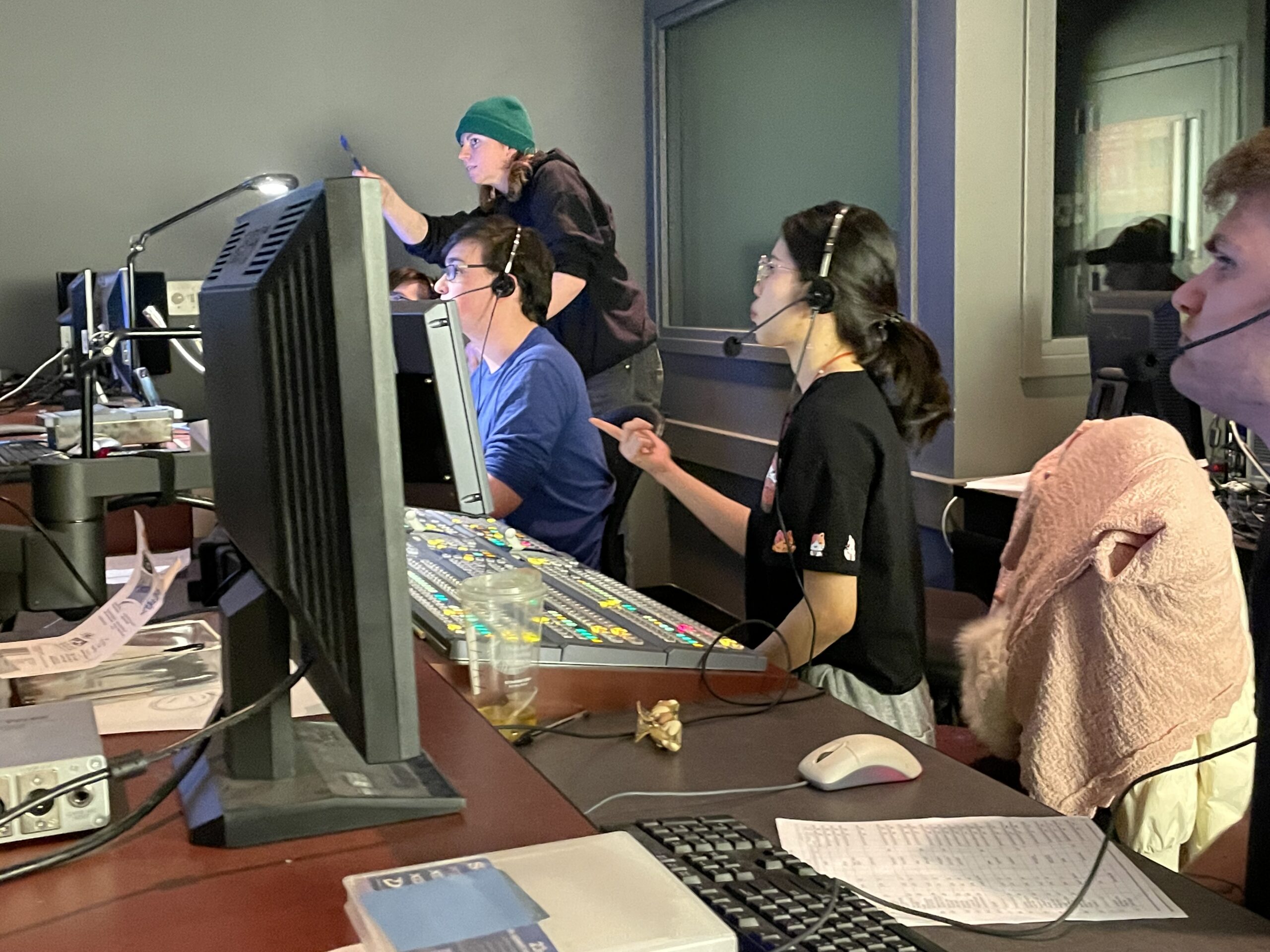 Several students wearing headsets work together in a television production control room. Multiple computer monitors, a video switcher and production equipment fill the workspace. One student stands behind the control stations pointing toward a monitor while others operate controls and review screens. Papers, a keyboard and other production materials are spread across the desks. The room is dimly lit, with the monitor glow illuminating the production team as they collaborate on a live broadcast or video production.