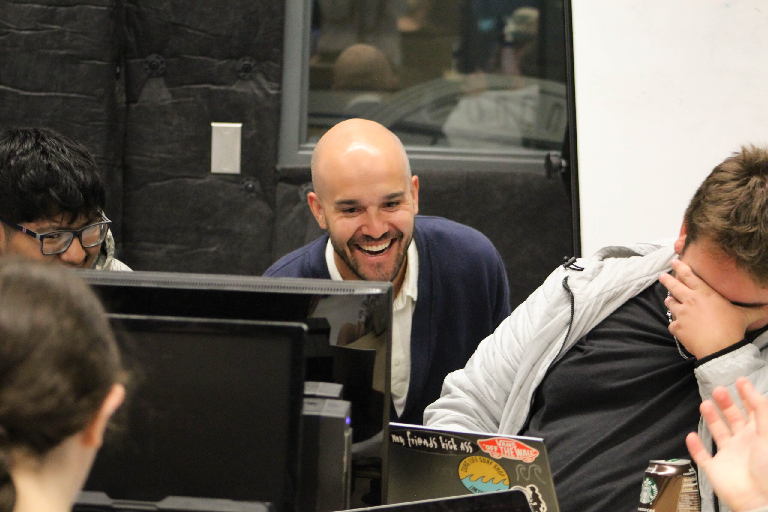 Students gather around computer workstations in a media or classroom lab. Several people are seated at desks with monitors and laptops, while a faculty member, Tony Thompson, leans toward the group during a discussion. A laptop covered with stickers is visible on the table, and a can of sparkling water sits nearby. The classroom features large interior windows and sound-dampening wall panels.