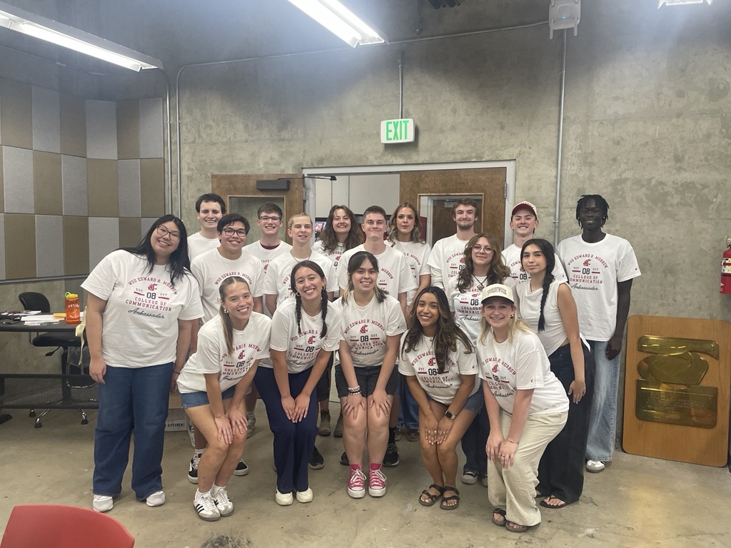 A group of approximately 15 students wearing matching Edward R. Murrow College of Communication T-shirts pose for a photo inside a media production or classroom space. The students stand in two rows in front of a concrete wall and double doors, with acoustic wall panels visible on the left. A large award plaque rests against the wall on the right side of the room. Tables, chairs and classroom equipment are visible in the background under bright overhead lighting.