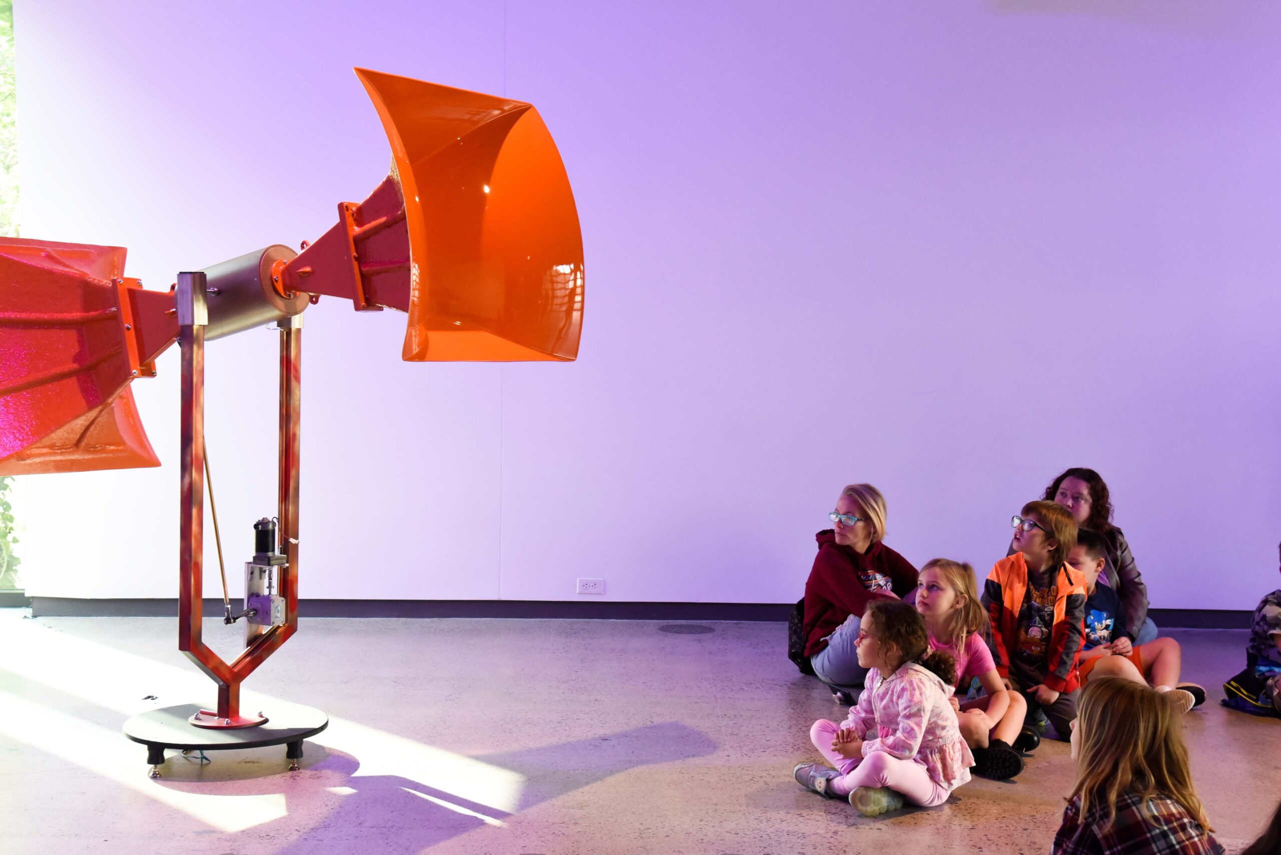 Children sit on the floor and observe an orange Trimpin horn. The wall is cast in purple light.