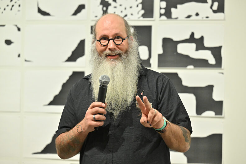 Squeak Meisel speaking into a microphone with black and white artwork in the background.