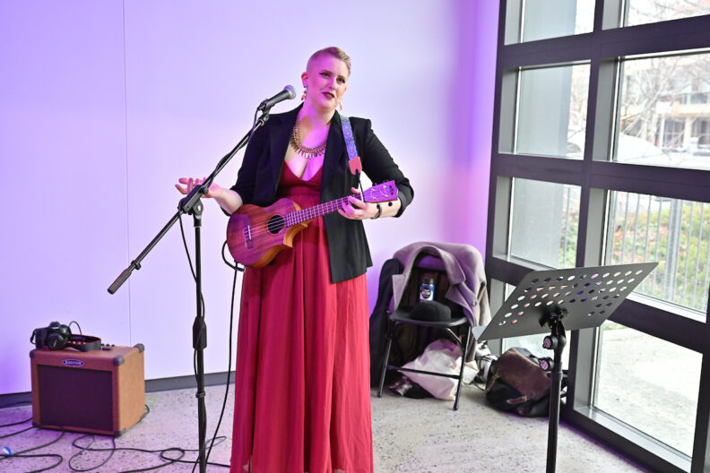 A musician with a ukulele speaking into a standing microphone in the pavilion gallery.