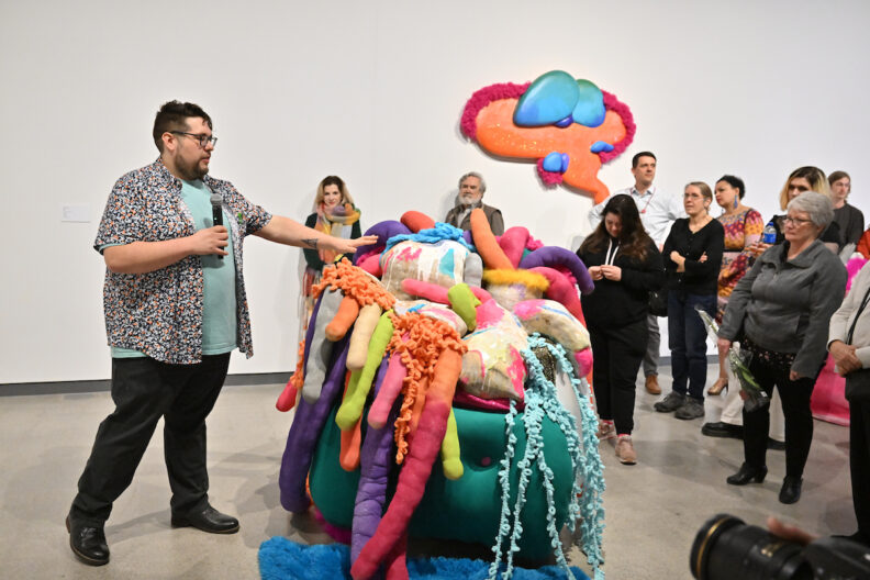 Adam Stuart gesturing towards one of his ceramic artworks in front of a crowd of people.