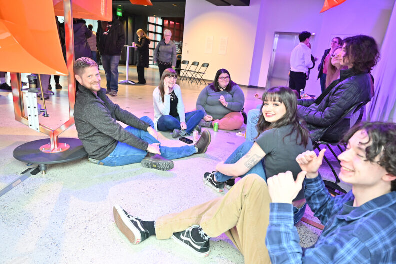 A group of visitors sitting on the pavilion gallery floor near an orange sculpture.