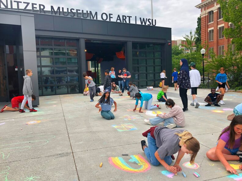 A group of fifth grade students creating chalk drawings on the ground in front of the art museum.