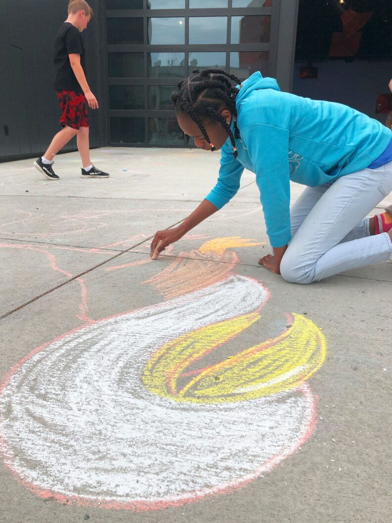 A fifth grade student creating a chalk drawing on the ground in front of the art museum.