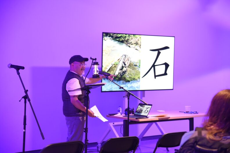 Sam Roxas-Chua speaks into a microphone and points at a presentation.