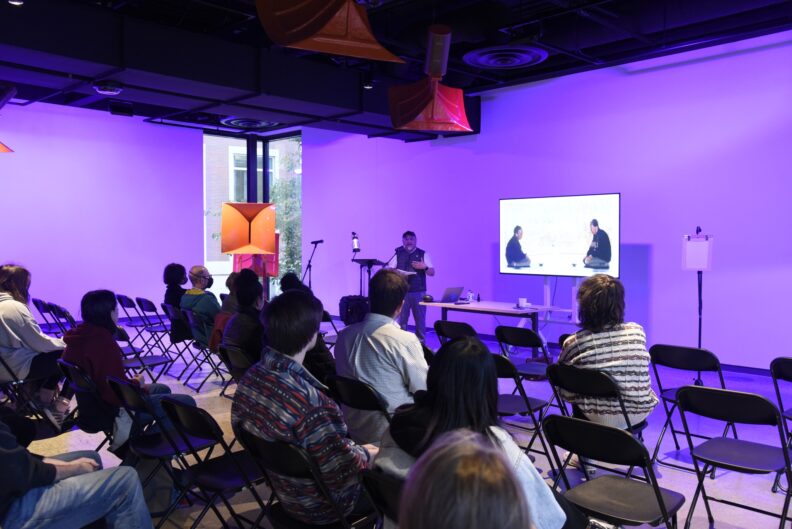 Sam Roxas-Chua speaks into a microphone in front of a seated group of people.