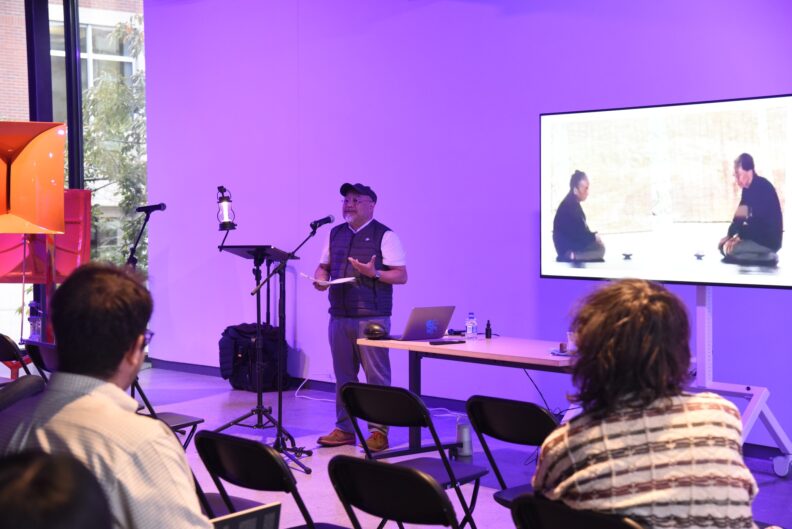 Sam Roxas-Chua speaks into a microphone next to a presentation.