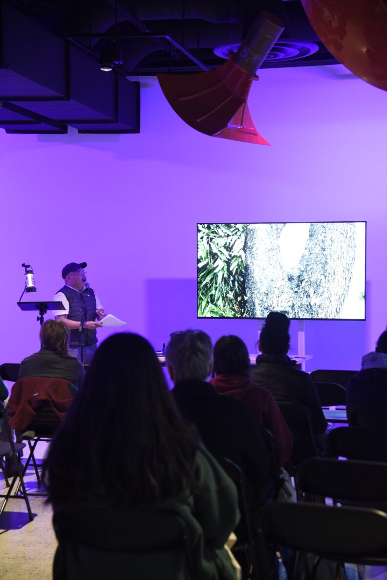 Sam Roxas-Chua speaks into a microphone next to a presentation showing a tree trunk and leaves.