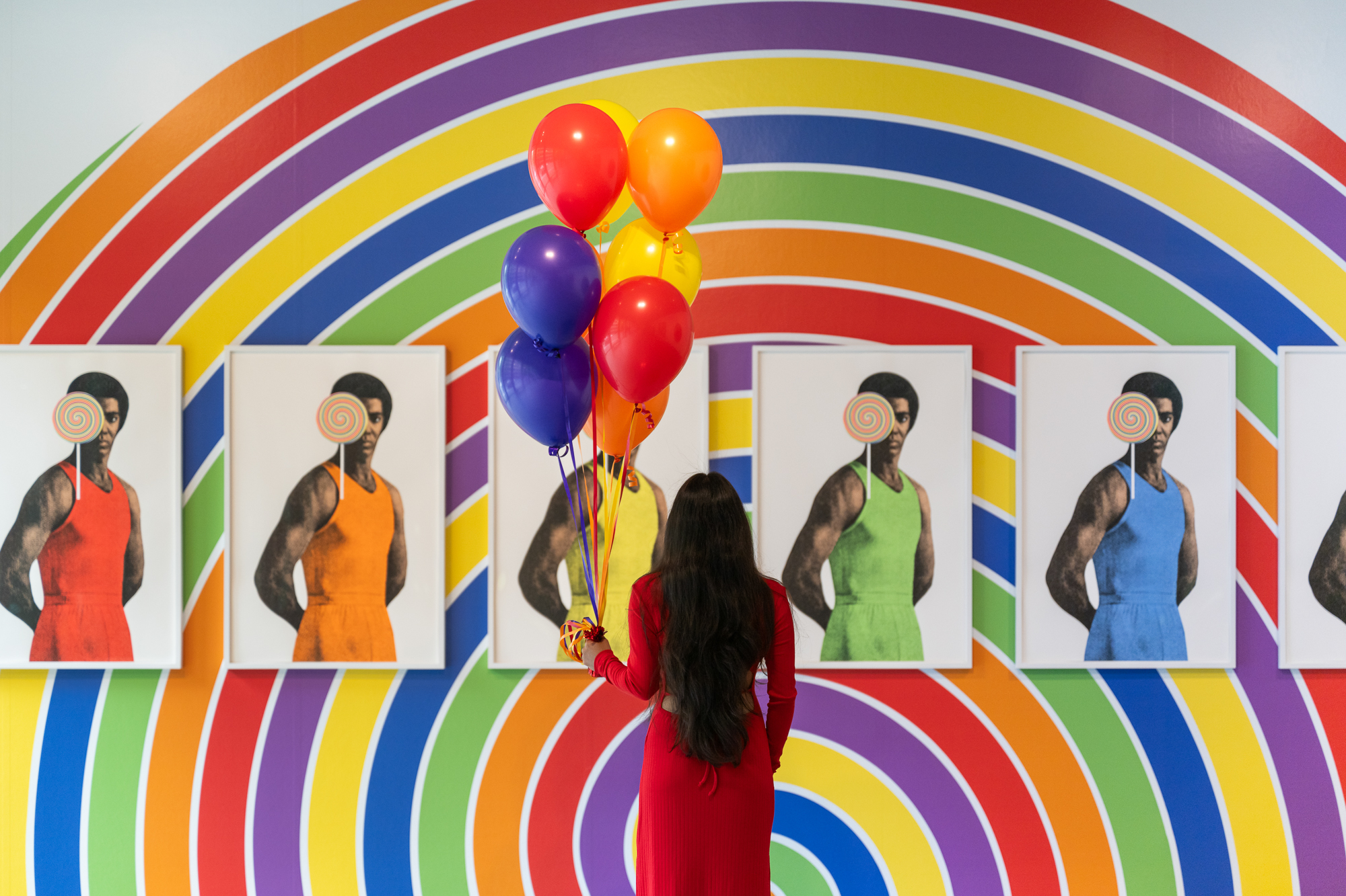Image of a woman in a red dress looking at a painted rainbow wall with photos and rainbow balloons.