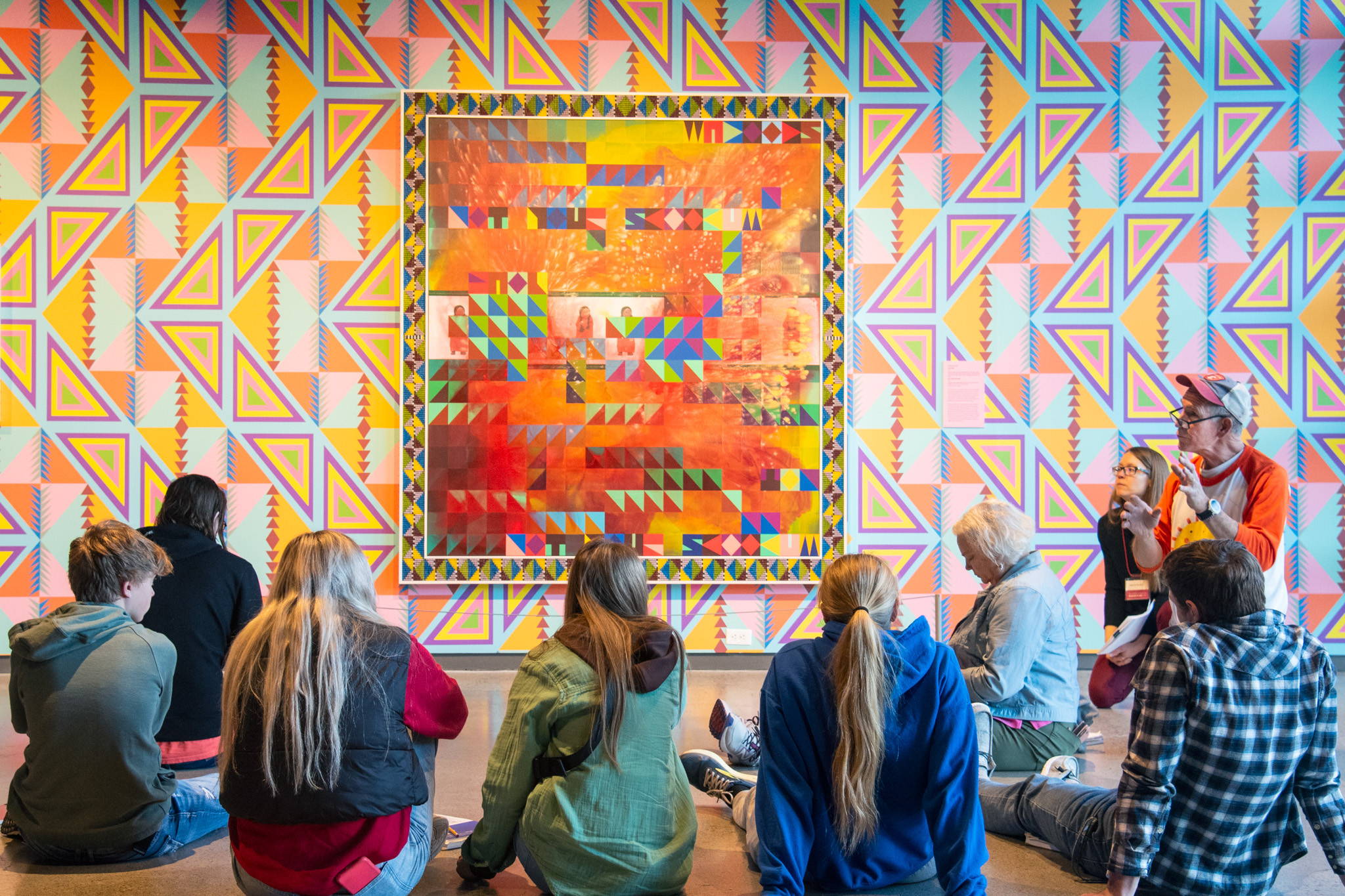 A class of students listens to a teacher while sitting in front of a colorful artwork.