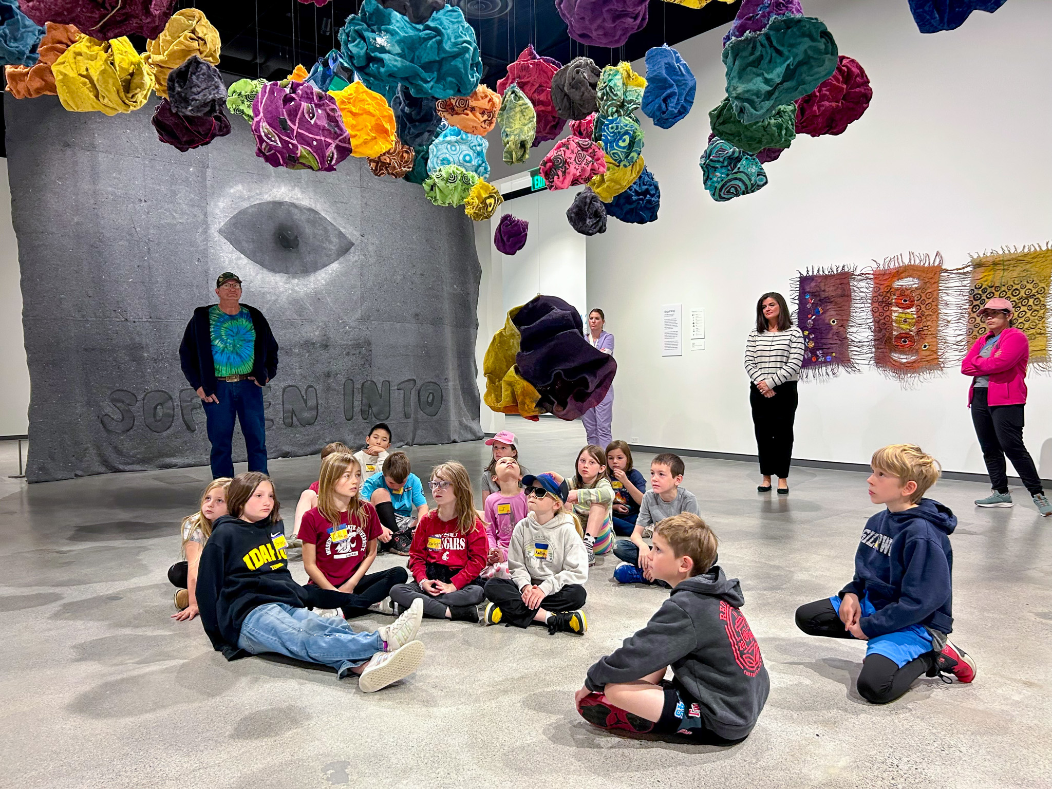 A group of elementary age children sits on the floor beneath a hanging art installation.