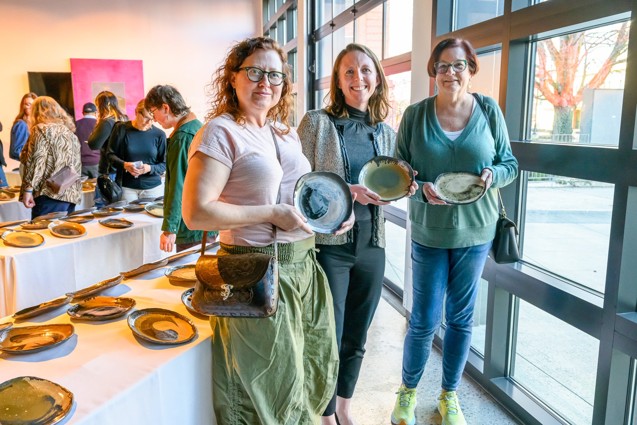 Three women smile and hold unique ceramic plates in the next to a window.